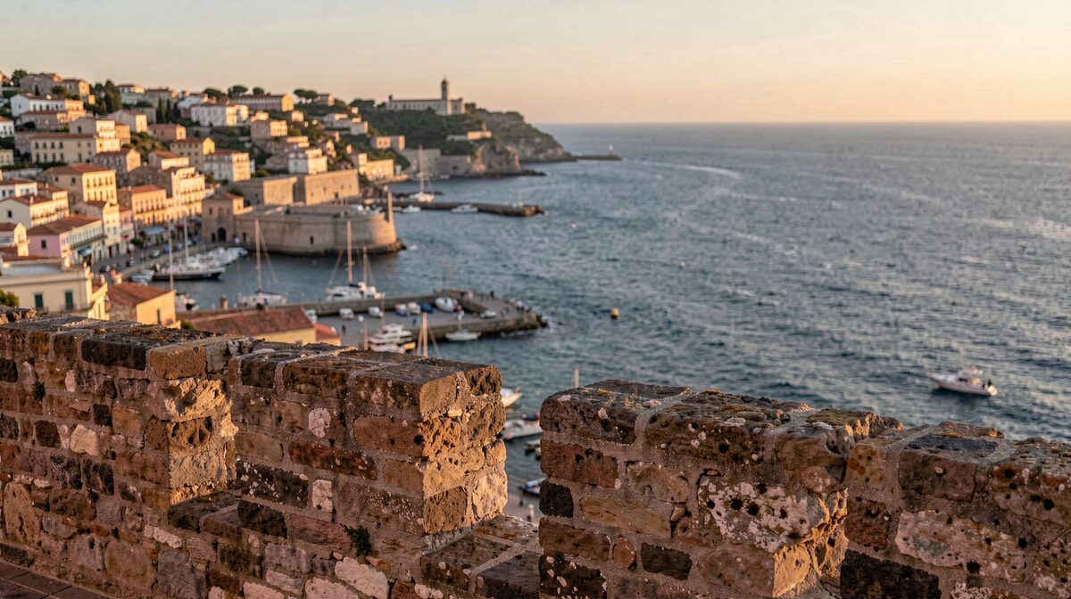Le soir sur les remparts d'Alghero, la lumière dorée touche les vieilles pierres et le port ; cadrage horizontal depuis les muralles vers la mer.
