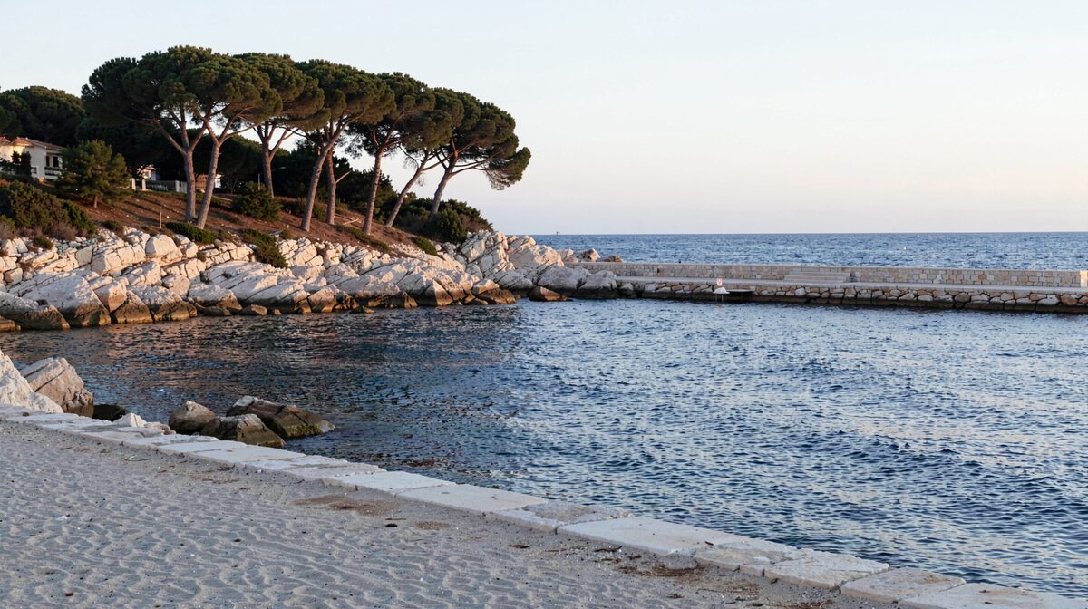 Une crique de galets et de sable clair encadrée par des pins parasols et un éperon rocheux blanc, un ponton en pierre avançant vers une mer calme en fin de journée, lumière dorée rasante, cadrage horizontal depuis le rivage.