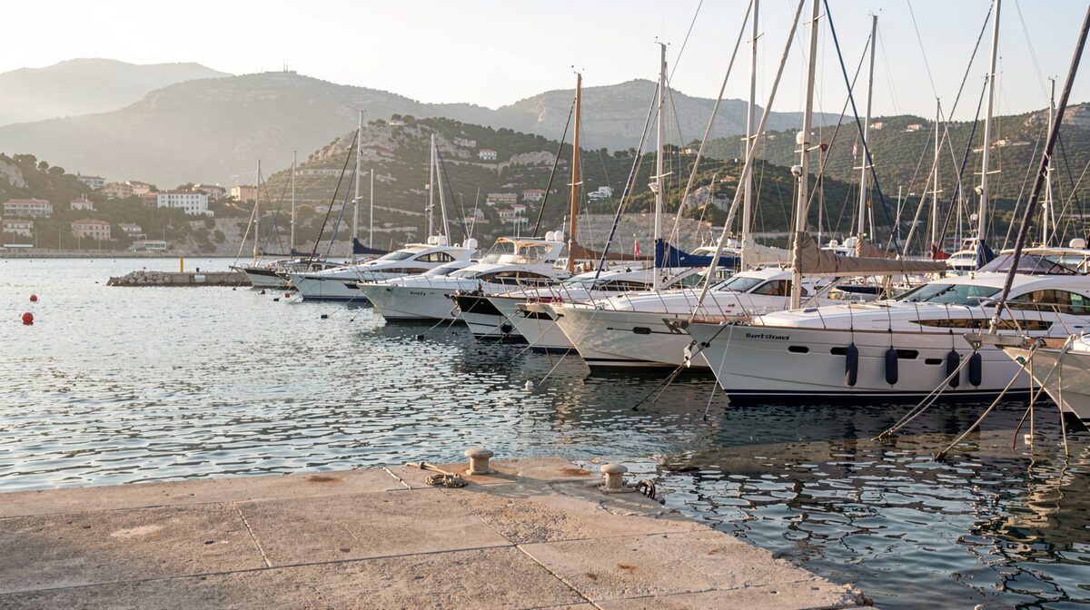 Le port de Saint-Tropez au petit matin, lumière rasante sur les coques alignées, un quai encore vide au premier plan et les collines du golfe en arrière-plan.